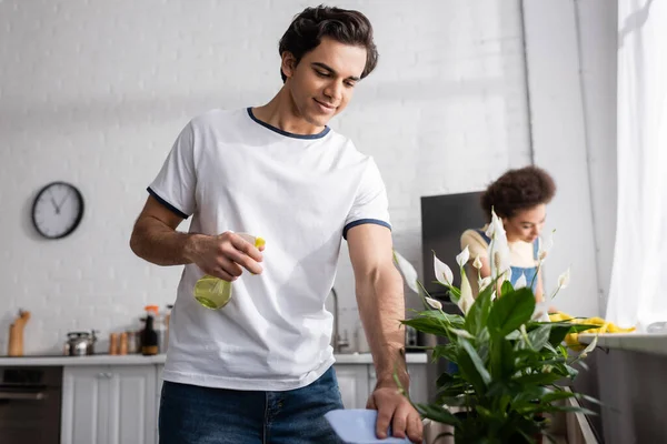 Smiling young man spraying water on plants near blurred african american girlfriend — Stock Photo