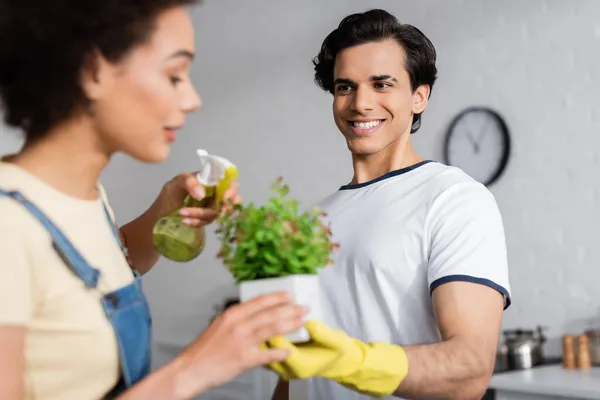 Happy young man holding plant near blurred african american woman with spray bottle — Stock Photo