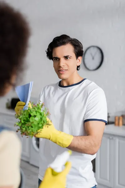 Joven hombre sosteniendo planta cerca borrosa africana americana mujer con aerosol botella - foto de stock