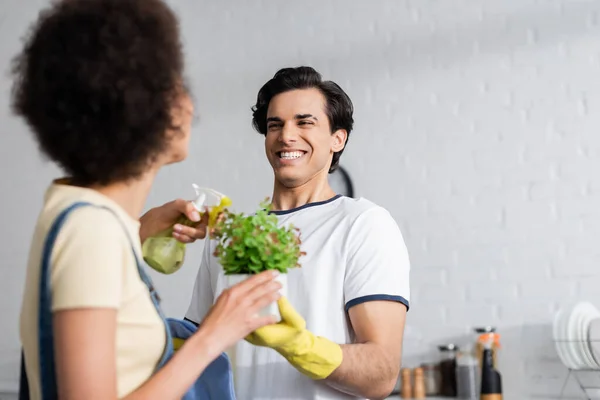 Happy young man holding green plant near blurred african american woman with spray bottle — Stock Photo