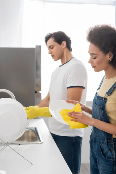 Curly african american woman cleaning plate with rag near blurred boyfriend — Stock Photo