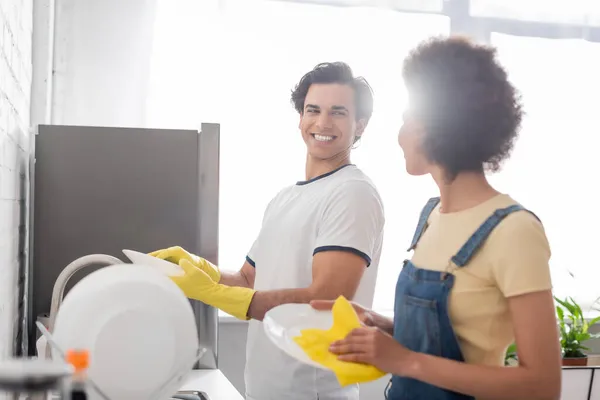 Glücklicher Mann spült Geschirr und schaut lockige Afroamerikanerin in der Küche an — Stockfoto