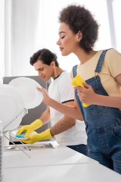 Smiling african american woman holding plate and rag near blurred boyfriend — Stock Photo