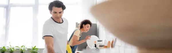 Jovem homem perto encaracolado afro-americano mulher segurando prato na cozinha, bandeira — Fotografia de Stock