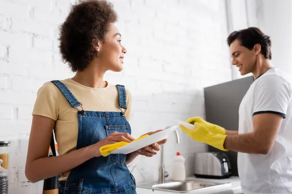 Lockige afrikanisch-amerikanische Frau lächelt, während sie Teller und Lappen in der Nähe verschwommener Freund beim Geschirrspülen hält — Stockfoto