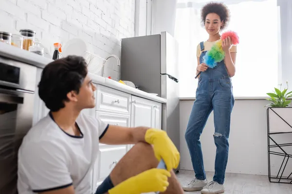 Curly african american woman holding dust brush and looking at blurred boyfriend in rubber gloves — Stock Photo