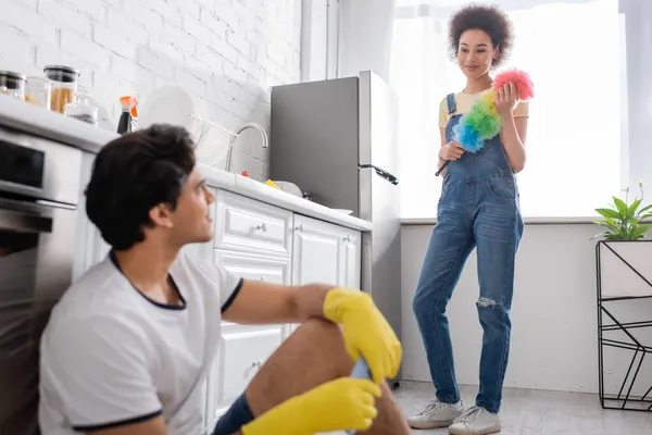 Lockige Afroamerikanerin hält Staubbürste in der Hand und schaut verschwommenen Freund in Gummihandschuhen an — Stockfoto