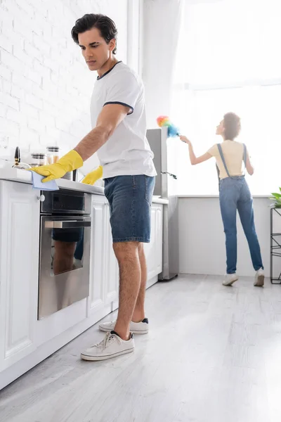 Молодий чоловік прибирає кухонний шафа з ганчіркою біля кучерявої афроамериканської жінки з пиловим пензлем на кухні — стокове фото