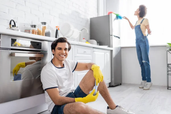 Jovem feliz sentado perto do armário da cozinha perto da mulher americana africana encaracolado limpeza geladeira com escova de poeira na cozinha — Fotografia de Stock