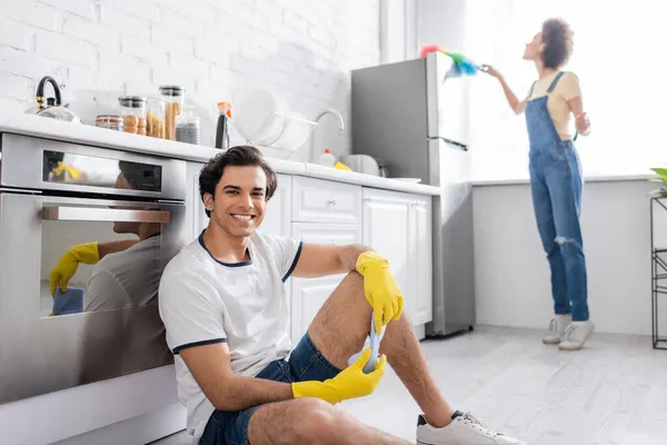 Feliz joven sentado cerca del gabinete de la cocina cerca de rizado mujer afroamericana limpieza nevera con cepillo de polvo en la cocina - foto de stock