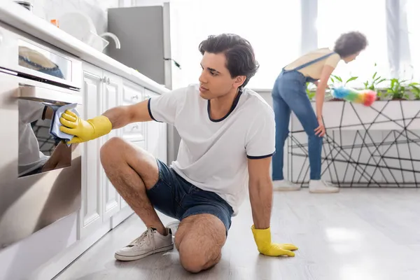 Hombre joven limpieza horno eléctrico con trapo cerca rizado mujer afroamericana con cepillo de polvo en la cocina - foto de stock