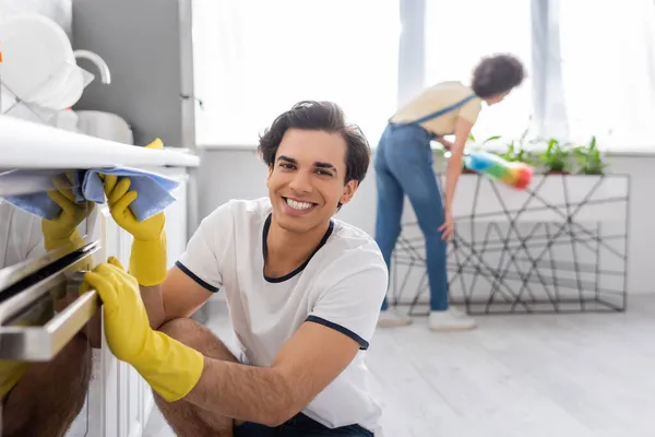 Feliz joven limpieza horno eléctrico con trapo cerca de rizado mujer afroamericana con cepillo de polvo en la cocina - foto de stock