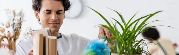 Young man doing house cleaning with dust brush near plants and books, banner — Stock Photo