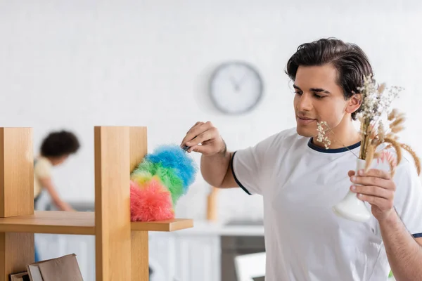 Young man cleaning wooden shelf with dust brush and holding plants in vase — Stock Photo