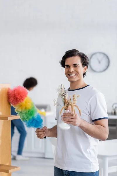 Heureux jeune homme nettoyage étagère en bois avec brosse à poussière et tenant des plantes dans un vase — Photo de stock