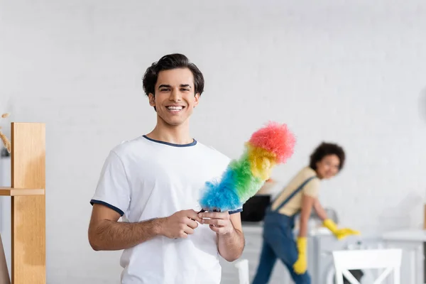 Happy young man holding dust brush near blurred african american woman — Stock Photo