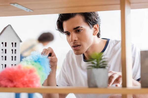 Young man cleaning wooden shelf with dust brush near plant — Stock Photo
