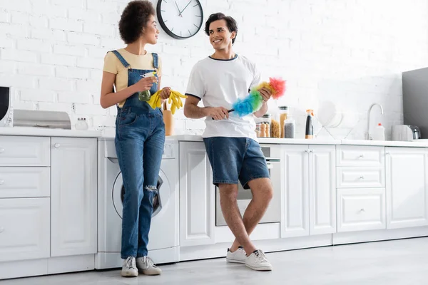 Longitud completa de joven feliz sosteniendo cepillo de polvo cerca de la novia afroamericana en guantes de goma - foto de stock