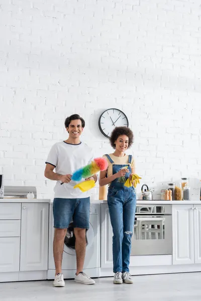 Full length of cheerful young man holding dust brush near african american girlfriend with rubber gloves — Stock Photo