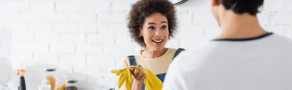 Mujer afroamericana rizada con guantes de goma mirando al hombre borroso en casa, pancarta - foto de stock