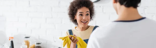 Encaracolado afro-americano mulher com luvas de borracha olhando para o homem borrado em casa, banner — Fotografia de Stock