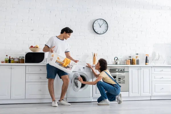 Happy african american woman looking at boyfriend holding basket with dirty laundry near washing machine in kitchen — Stock Photo