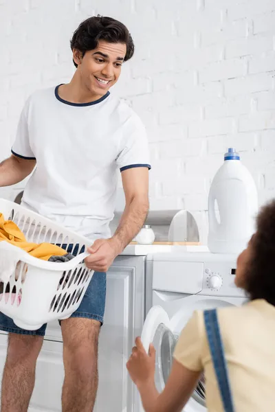 Homme heureux tenant panier avec sale buanderie près de la machine à laver et floue petite amie afro-américaine — Photo de stock