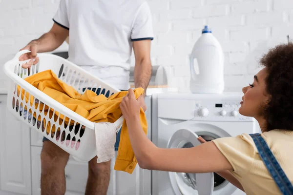 Encaracolado afro-americano mulher olhando para cesta com roupa suja perto máquina de lavar roupa e namorado — Fotografia de Stock