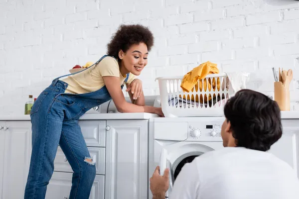 Glücklich afrikanisch-amerikanische Frau schaut Freund in der Nähe Korb mit schmutziger Wäsche auf Waschmaschine — Stockfoto