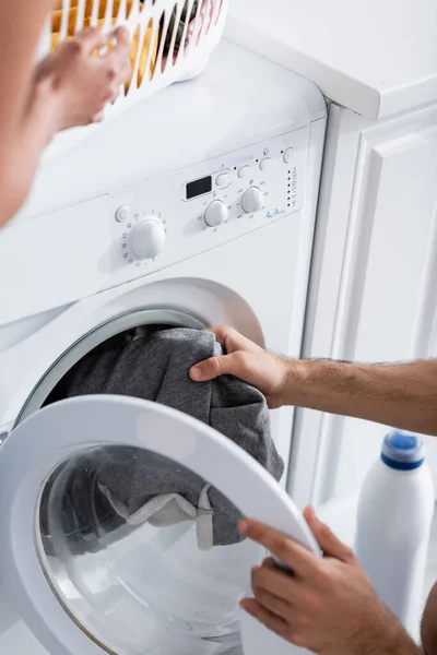 Cropped view of man holding laundry near washing machine and blurred african american girlfriend — Stock Photo