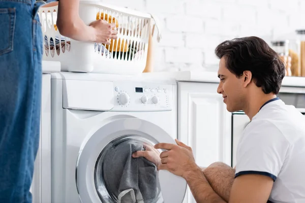 Улыбающийся мужчина погрузки стиральная машина рядом с африканской американской подругой с корзиной грязного белья — стоковое фото