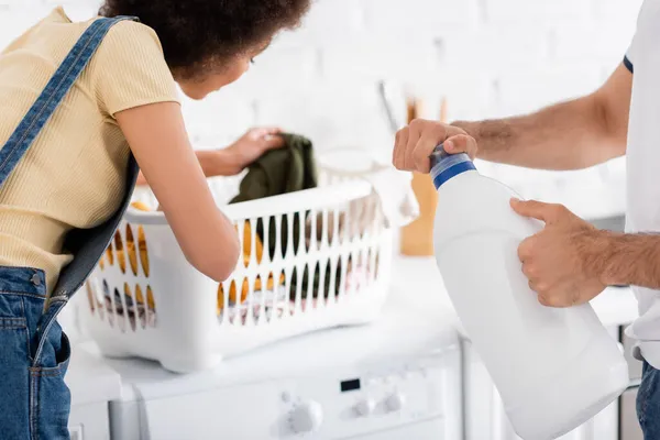 Homem segurando garrafa com detergente perto encaracolado afro-americano namorada olhando para cesta de roupa suja — Fotografia de Stock