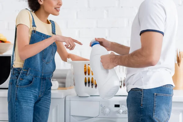 Vista recortada de mujer afroamericana apuntando a botella con detergente cerca de novio y cesta de ropa sucia - foto de stock