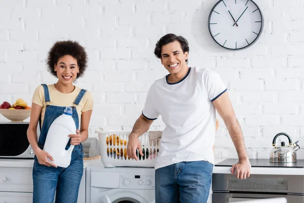 Happy african american woman holding bottle with detergent near boyfriend and basket of dirty laundry — Stock Photo