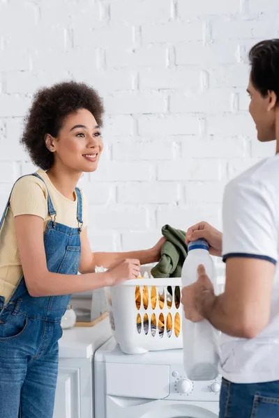 Mujer afroamericana feliz mirando novio sosteniendo botella con detergente - foto de stock