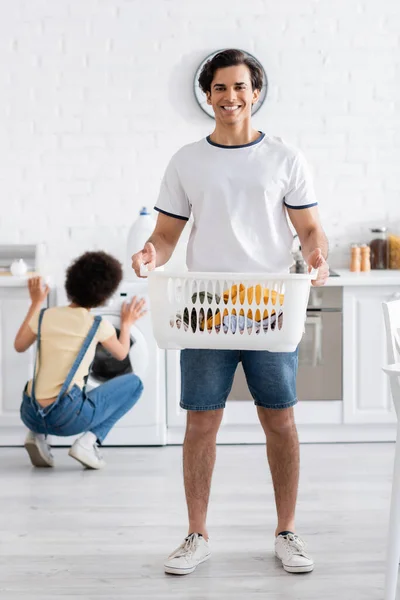 Cheerful man holding basket with dirty laundry near african american woman — Stock Photo