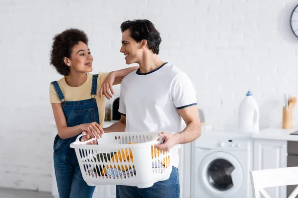Glückliches gemischtrassiges Paar schaut einander in der Nähe von Korb mit schmutziger Wäsche an — Stockfoto
