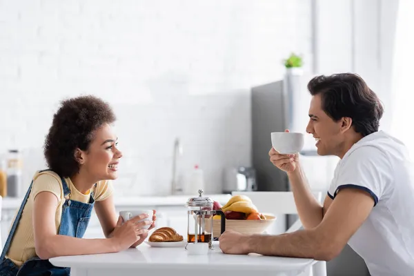 Glückliches interrassisches Paar lächelt, während es sich beim Frühstück ansieht — Stockfoto