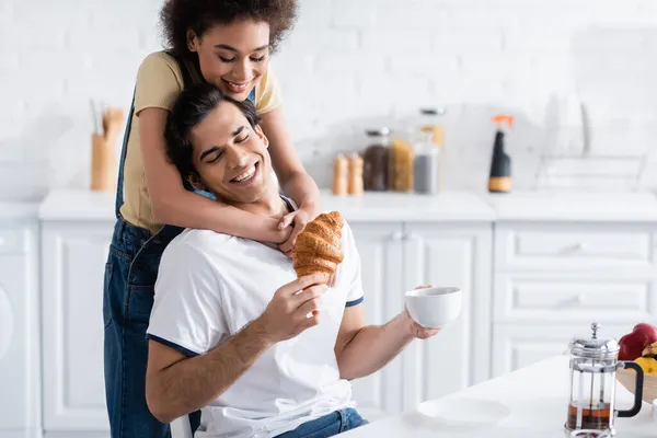 Feliz afro-americano mulher abraçando namorado com xícara de chá e croissant — Fotografia de Stock