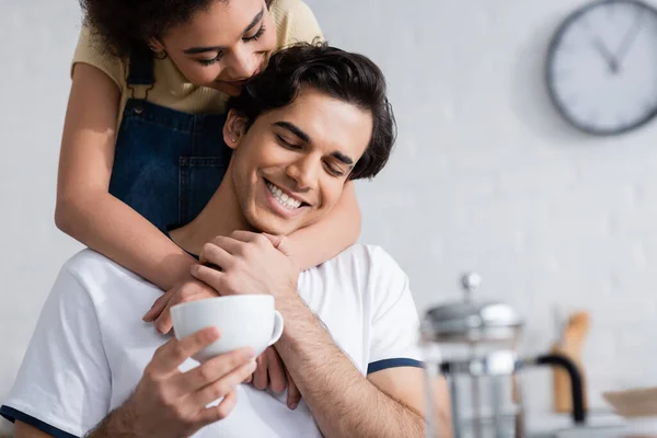 Heureuse femme afro-américaine étreignant petit ami avec tasse de thé dans la cuisine — Photo de stock