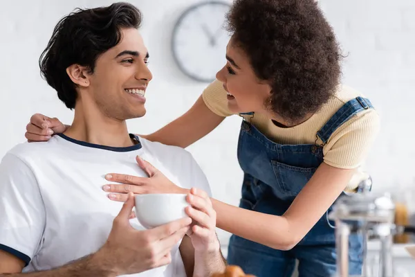 Smiling african american woman hugging boyfriend with cup of tea in kitchen — Stock Photo