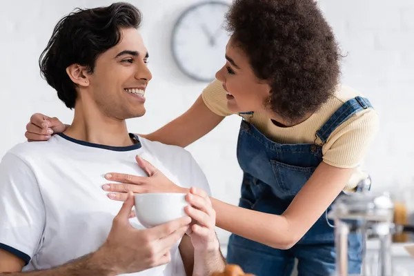 Sonriente africano americano mujer abrazando novio con taza de té en cocina - foto de stock