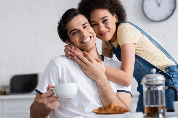 Gioiosa donna afroamericana che abbraccia il fidanzato con una tazza di tè in cucina — Foto stock