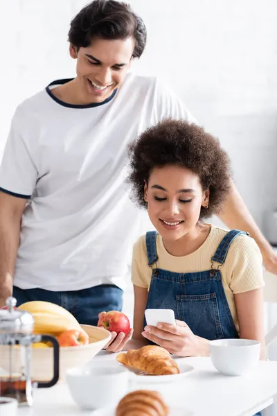 Glückliches gemischtes Paar schaut beim Frühstück aufs Smartphone — Stockfoto