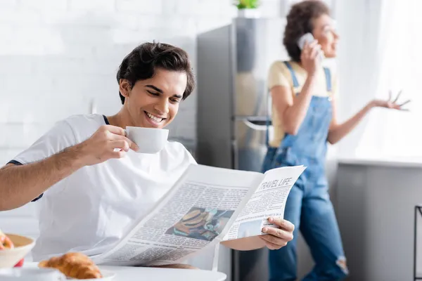 Cheerful man holding cup and reading newspaper while blurred african american woman talking on smartphone — Stock Photo