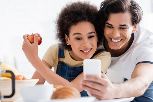 Fröhliches gemischtes Paar schaut während des Frühstücks aufs Smartphone — Stockfoto