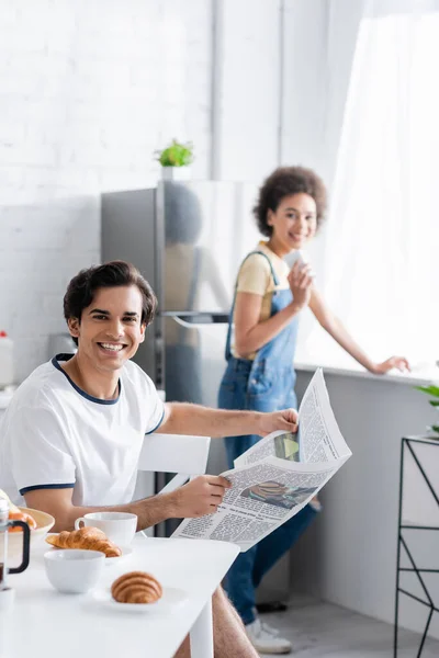 Cheerful man holding newspaper near blurred african american woman with smartphone — Stock Photo