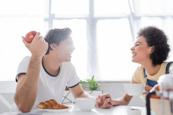 Vista laterale della felice coppia multietnica che si tiene per mano durante la colazione — Foto stock