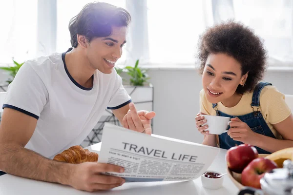 Happy man pointing at travel life newspaper near african american girlfriend during breakfast — Stock Photo