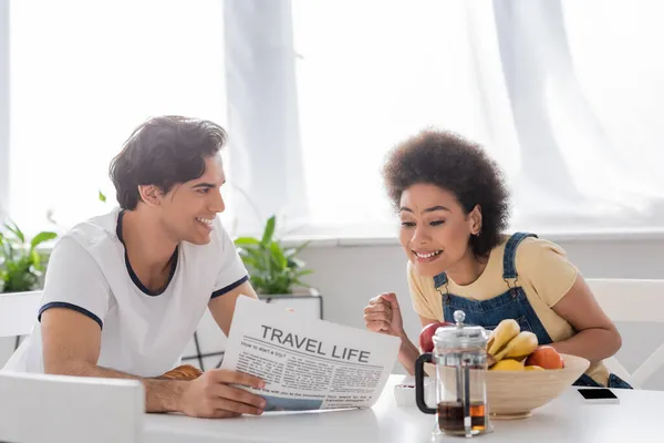 Happy man holding travel life newspaper near african american girlfriend during breakfast — Stock Photo